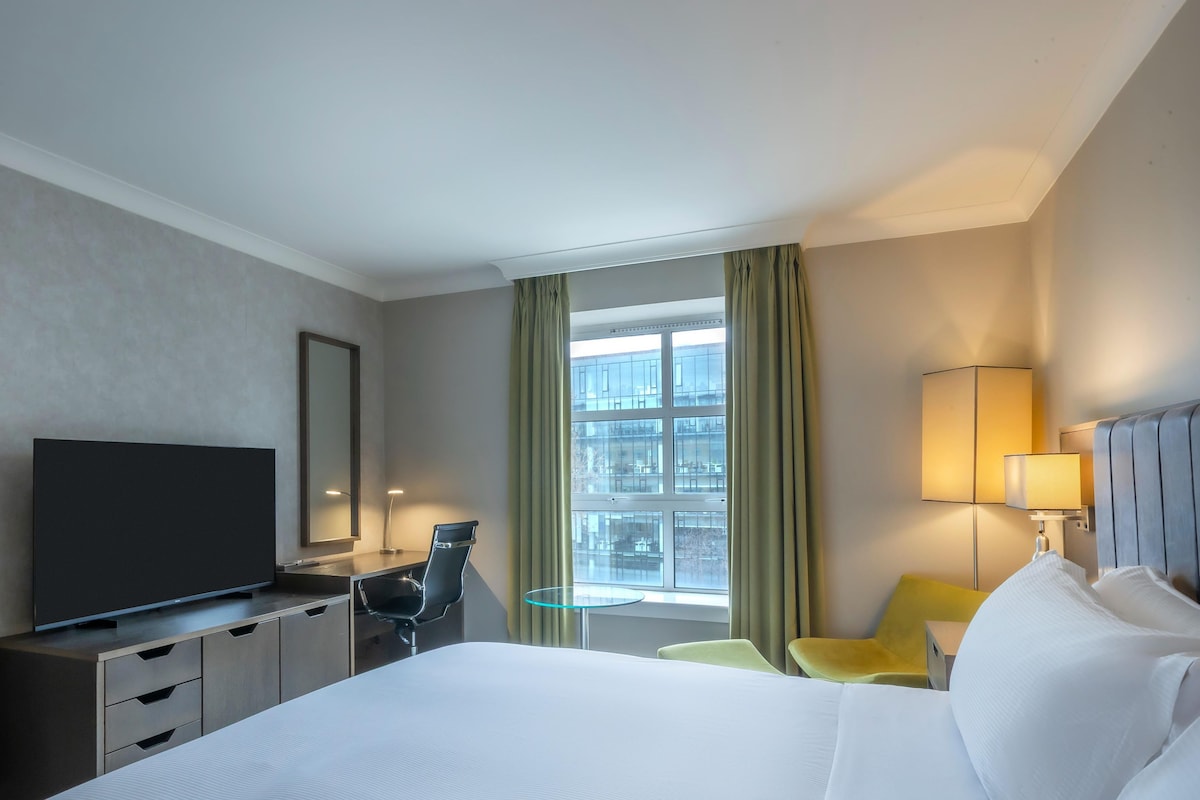 Une chambre d’hôtel équipée d’un lit, d’un bureau, d’une télévision, d’une chaise, d’une lampe et d’une fenêtre avec des rideaux.