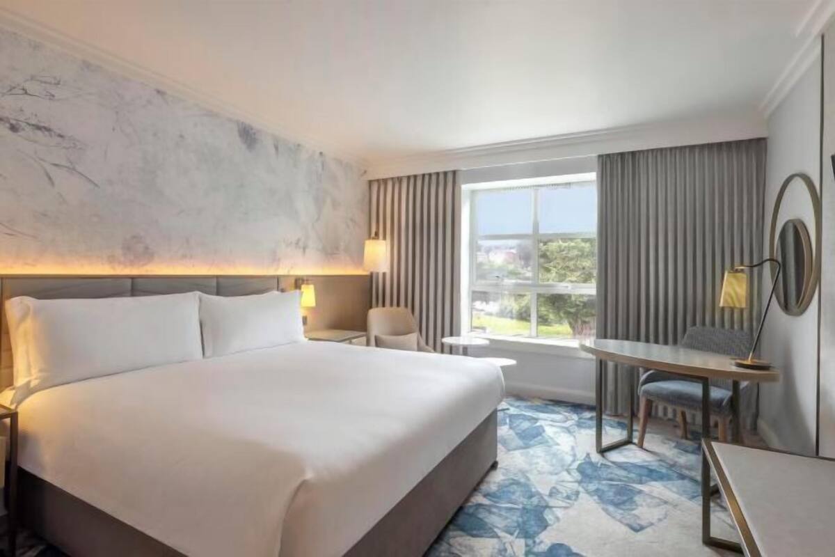 Habitación de hotel con una cama grande, un escritorio, una silla, una lámpara y vistas al exterior.