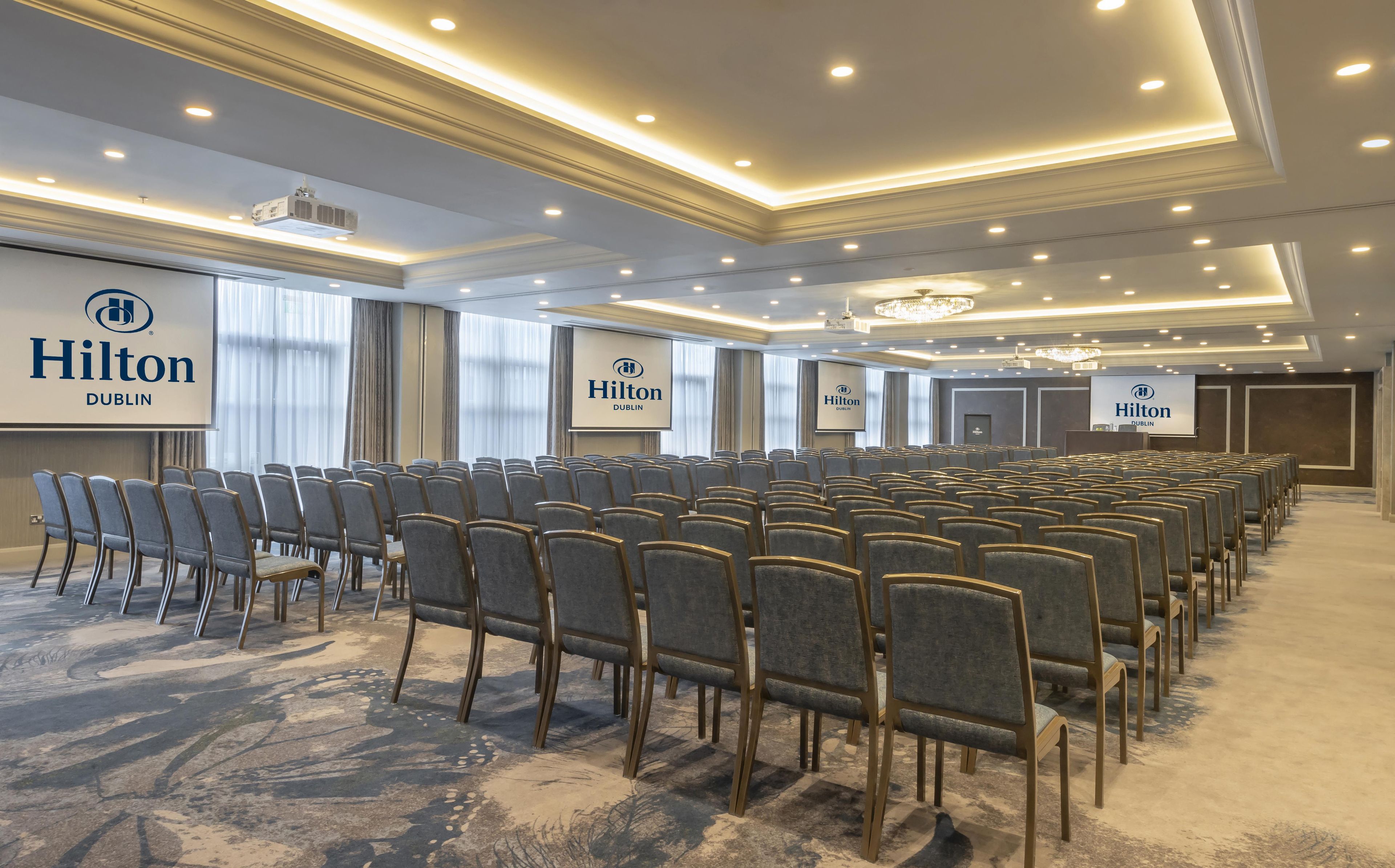 A conference room with rows of chairs, a projector screen, and a large screen displaying the Hilton Dublin logo.