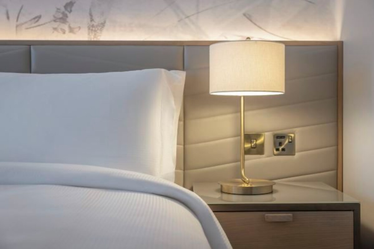 A neatly made bed with a white comforter and pillows, a bedside table with a lamp, and a wall-mounted control panel.