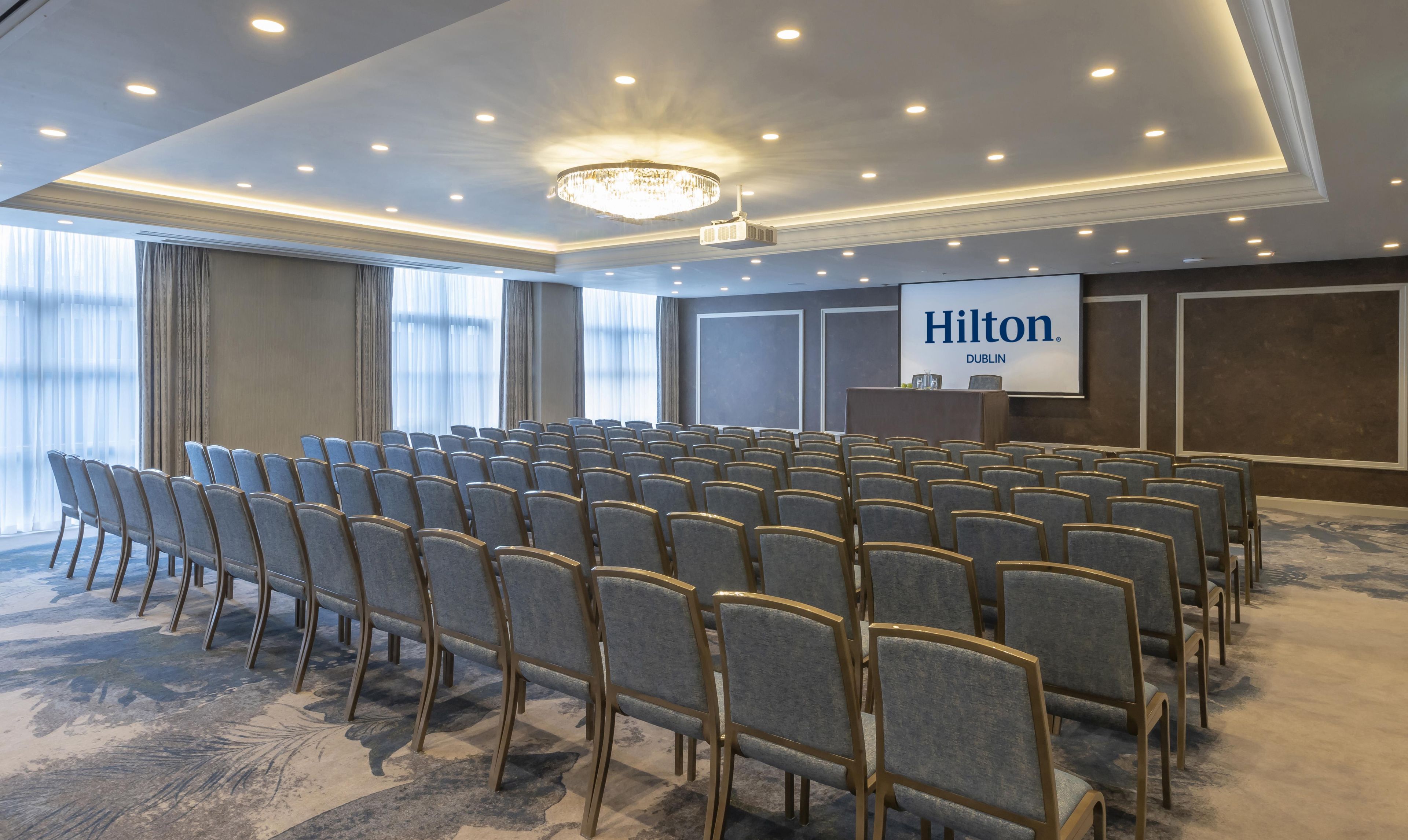 Une salle de conférence avec des rangées de chaises, un pupitre et un écran affichant le texte « Hilton Dublin ».