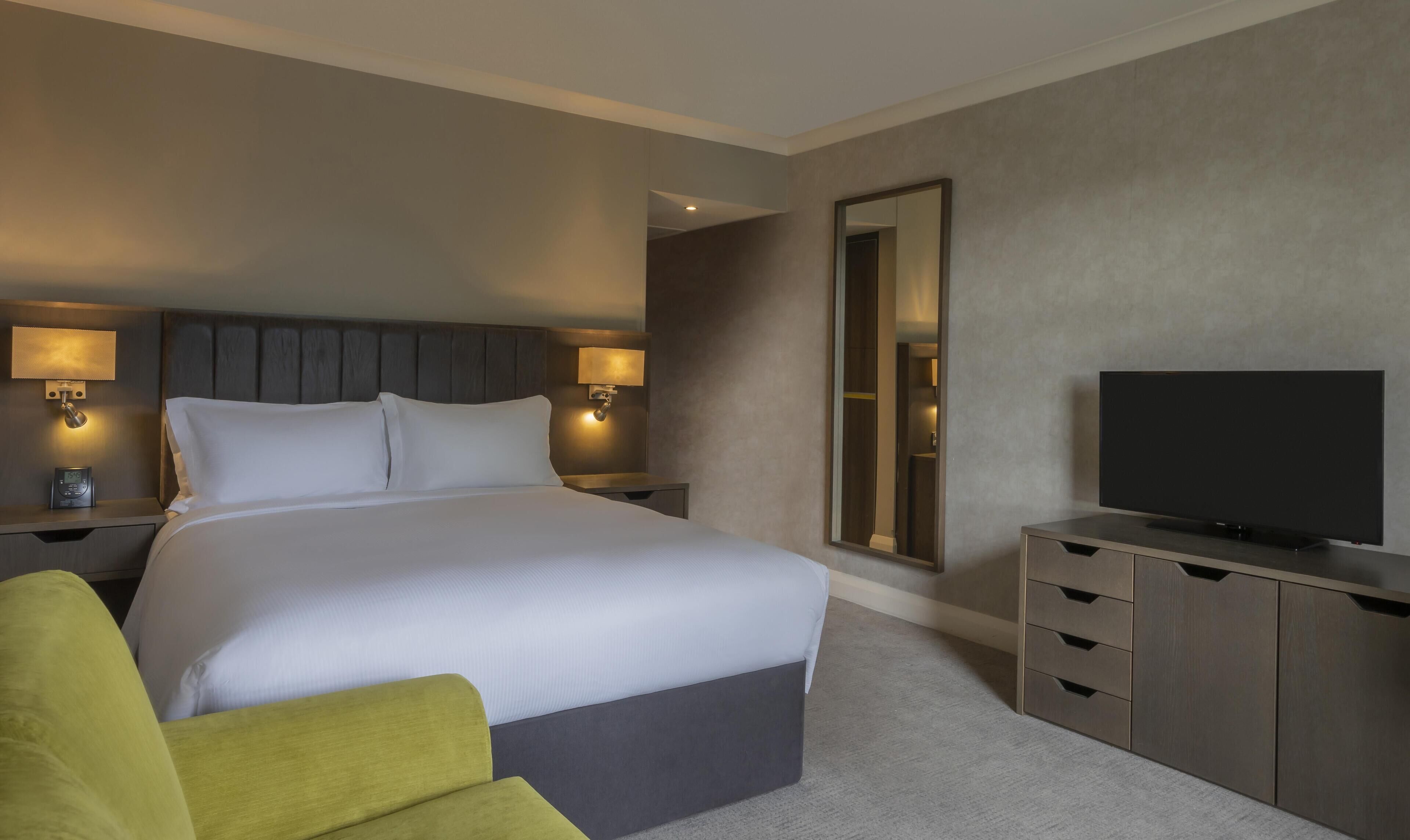 A hotel room with a large bed, bedside lamps, a flat-screen TV, a mirror, and a small seating area.