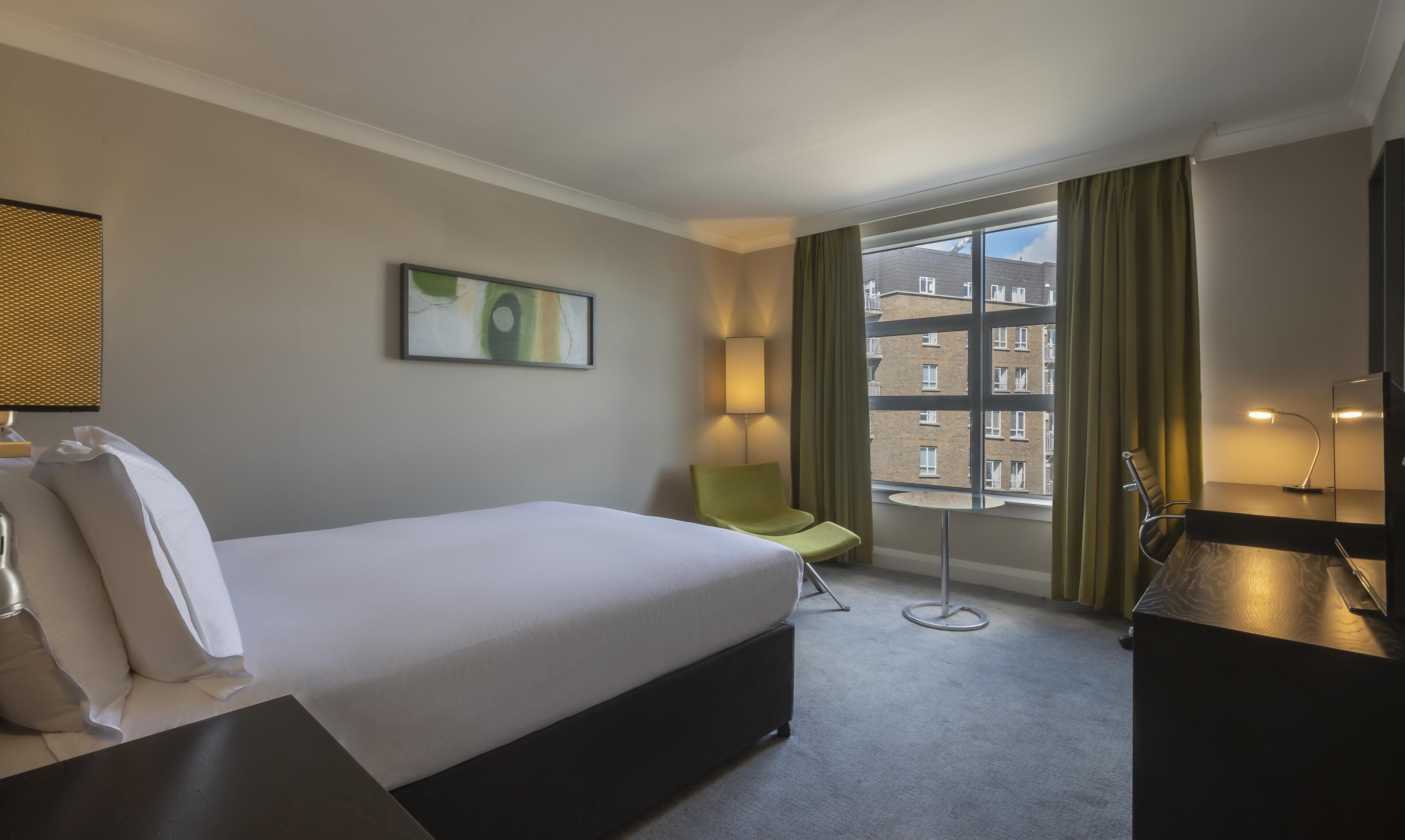 Habitació d'hotel amb llit, una cadira, escriptori, làmpada, quadre i finestra amb cortines.