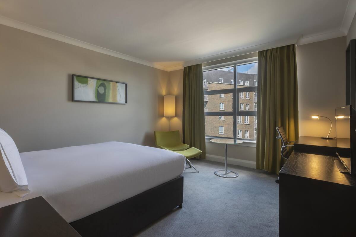 Habitación de hotel con cama, una silla, escritorio, lámpara, un cuadro y ventana con cortinas.