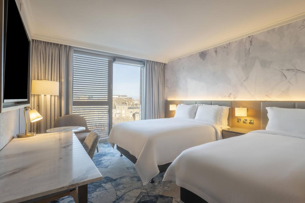 Habitación de hotel con dos camas, un escritorio, una silla y un ventanal con persianas.