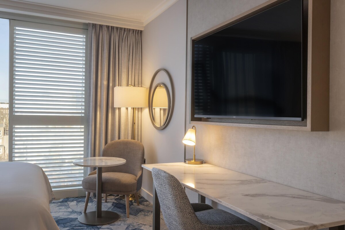 Une chambre d’hôtel avec un lit, une table ronde, une chaise, un bureau, une lampe, un miroir et une grande fenêtre avec des stores.