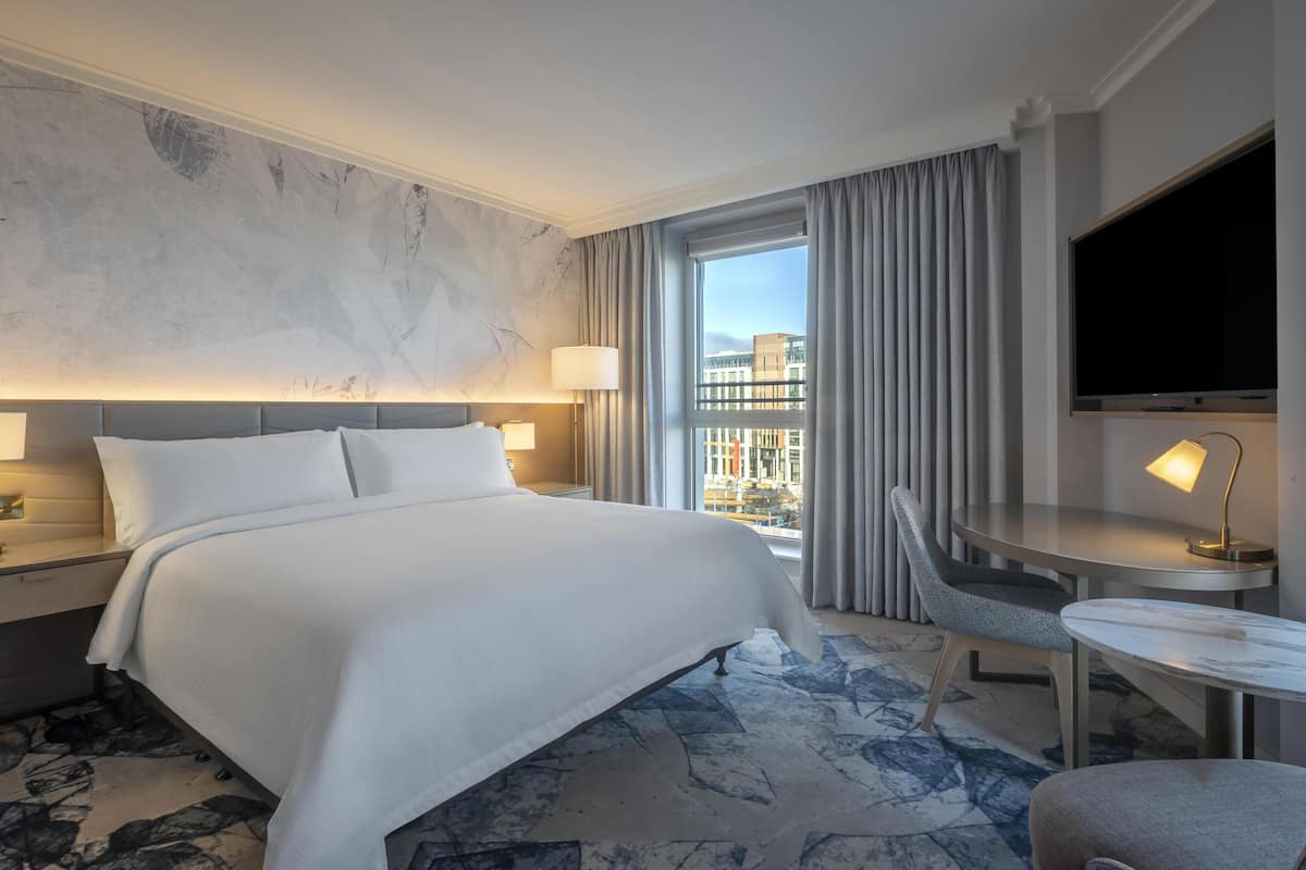 A hotel room with a large bed, a desk with a chair, a TV, and a view of buildings through the window.