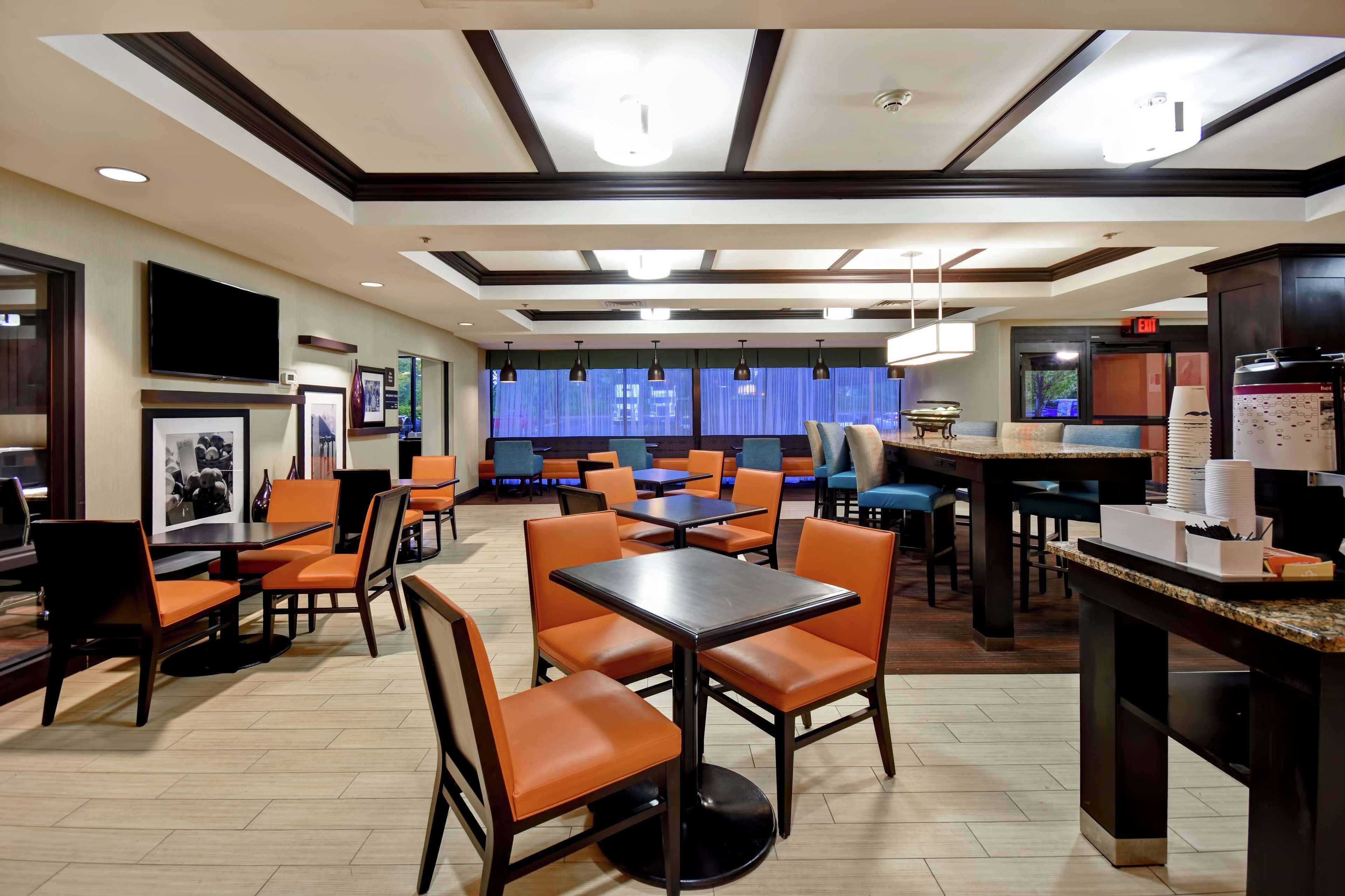 A hotel dining area with multiple tables and orange chairs, a flat-screen TV mounted on the wall, and a ceiling with recessed lighting.