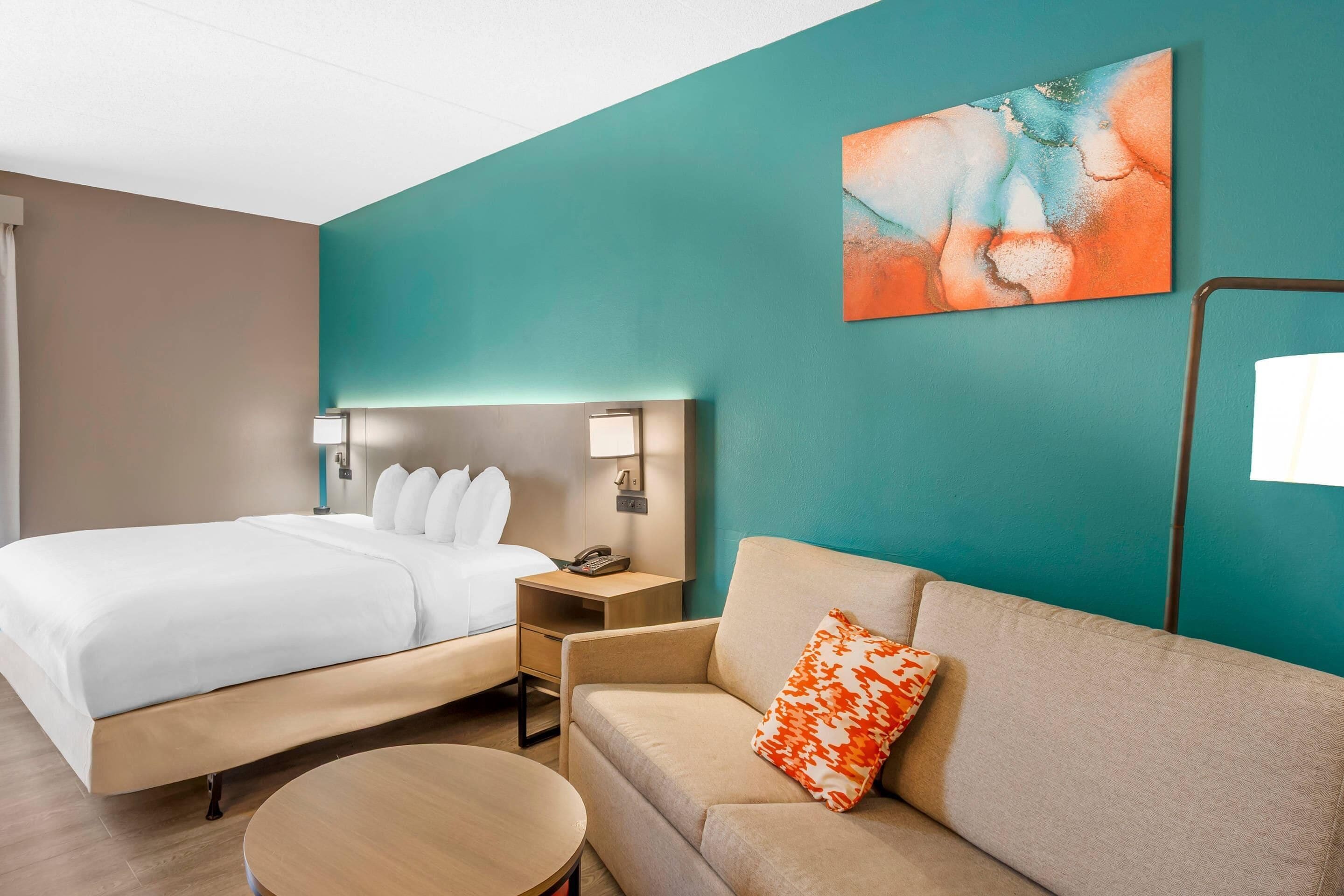 Una habitación de hotel con cama, sofá, mesita de noche, lámpara y un cuadro en la pared.