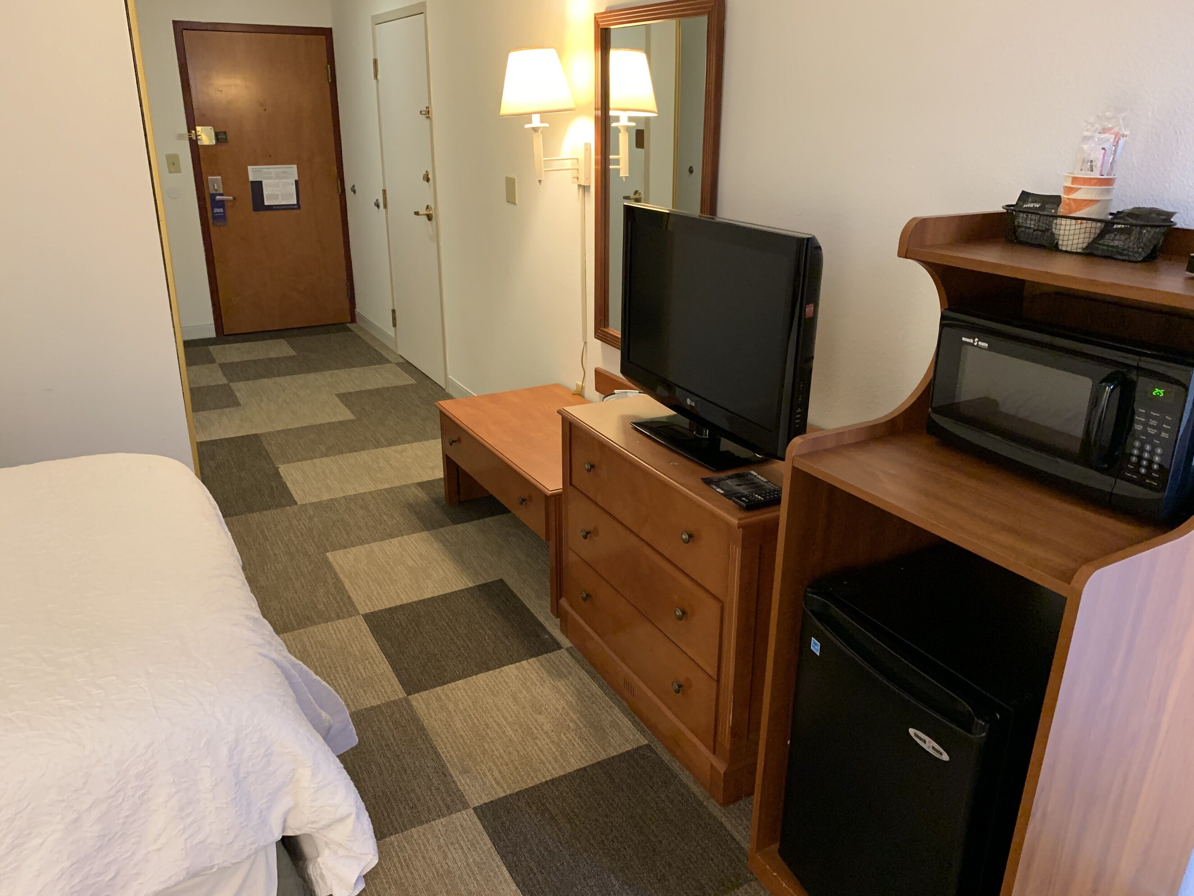 Habitación de hotel con cama, televisión, microondas, refrigerador pequeño, cómoda de madera y escritorio con lámpara.