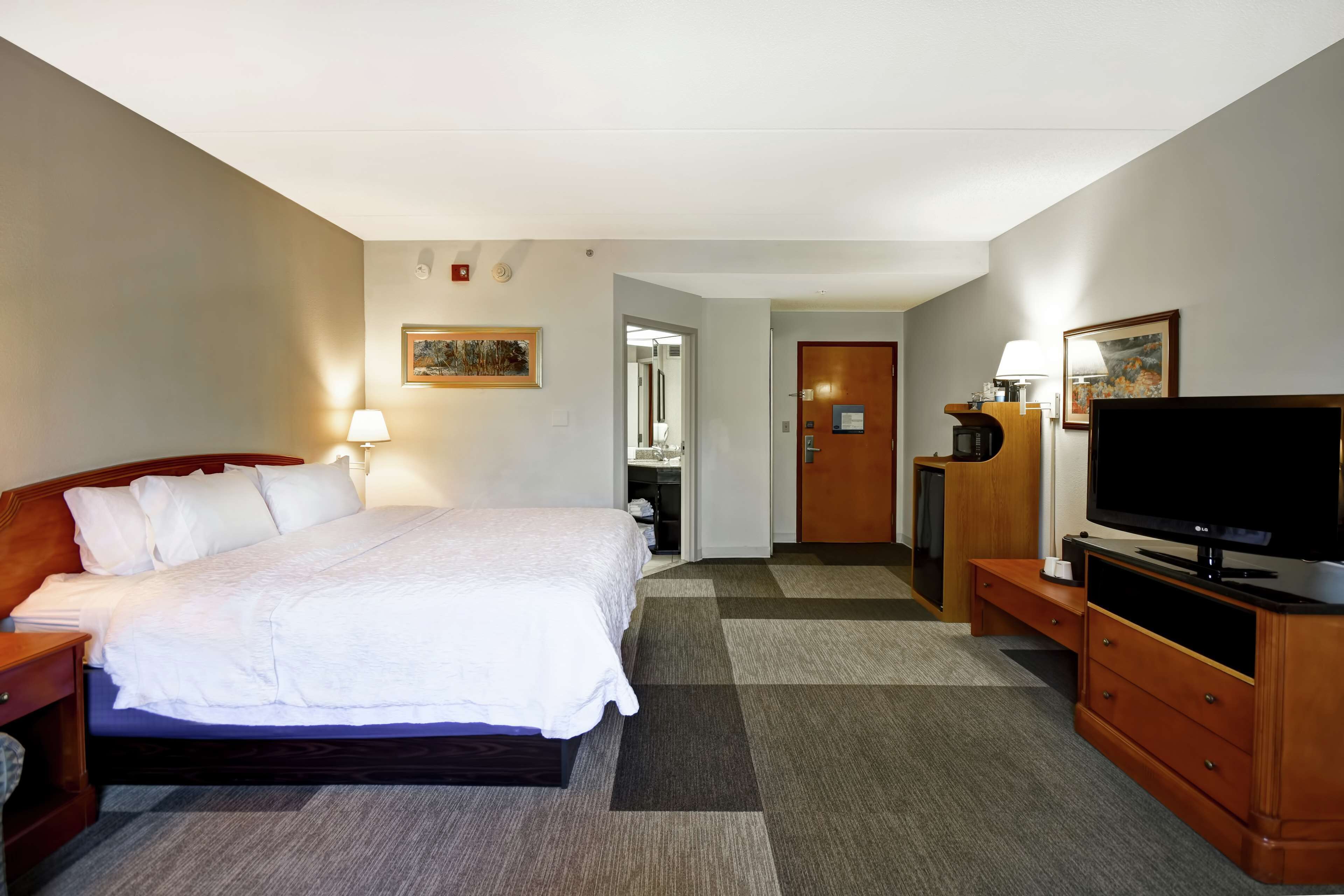 A hotel room with a bed, bedside table, lamp, TV, and a wooden dresser.