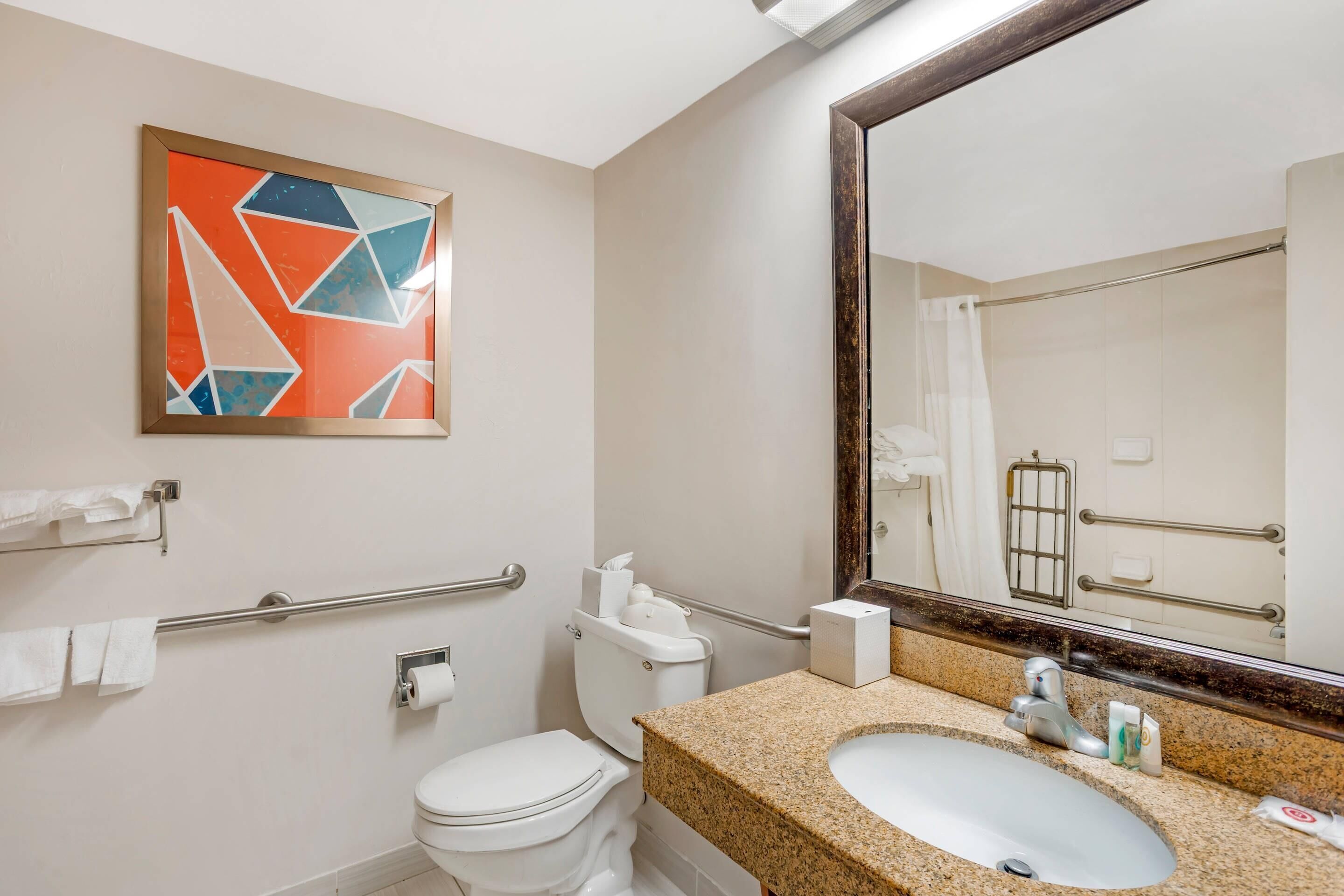 Une salle de bain moderne avec des toilettes blanches, un lavabo doté d’un plan de travail en granit, un grand miroir et une barre d’appui pour faciliter l’accès.