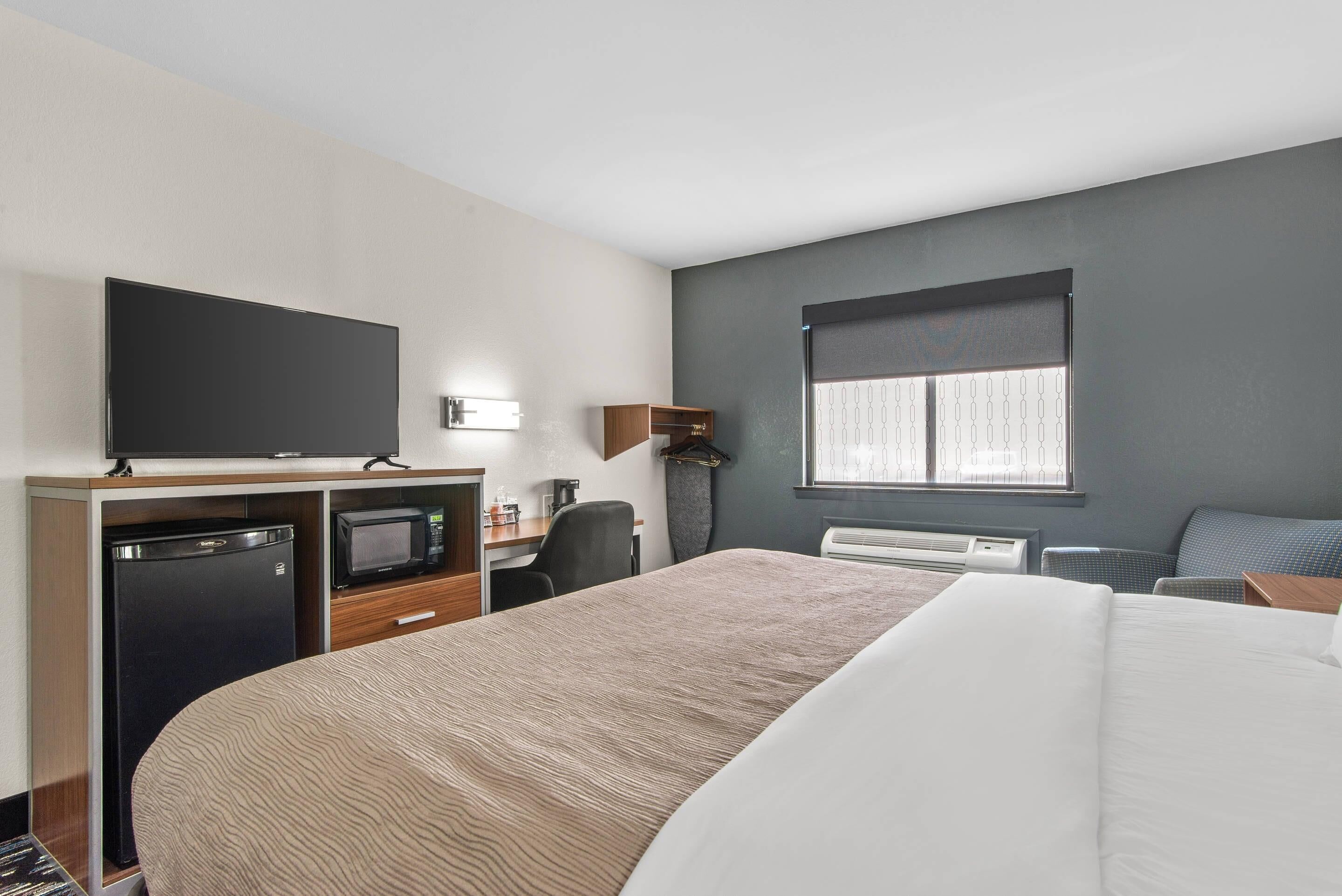 A hotel room with a bed, a desk with a TV, a microwave, a mini-fridge, and a window with blinds.