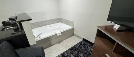 A modern hotel room with a sofa, a TV, a bathtub, and a desk with a sink.