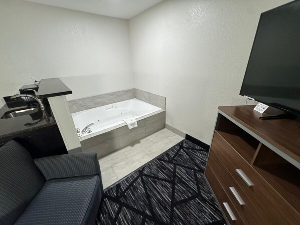 A modern hotel room with a sofa, a TV, a bathtub, and a desk with a sink.