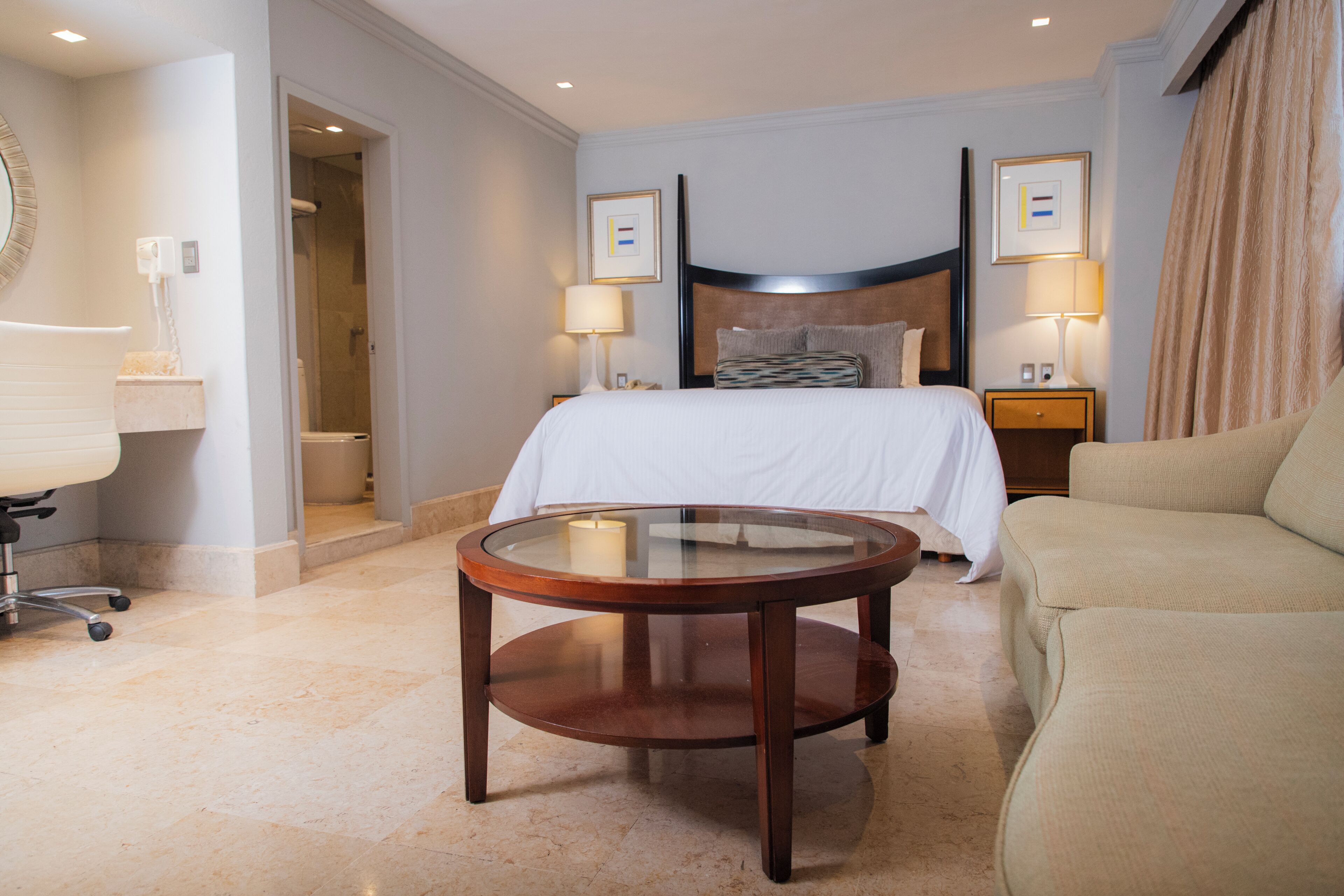 A hotel room with a bed, two bedside lamps, a round wooden table, a sofa, a bathroom with a shower, and a framed picture on the wall.