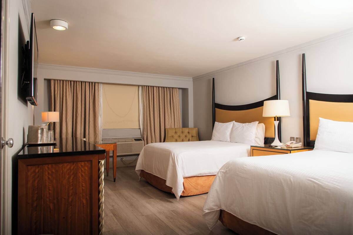 A hotel room with two beds, a desk, a chair, a TV, and a window with curtains.