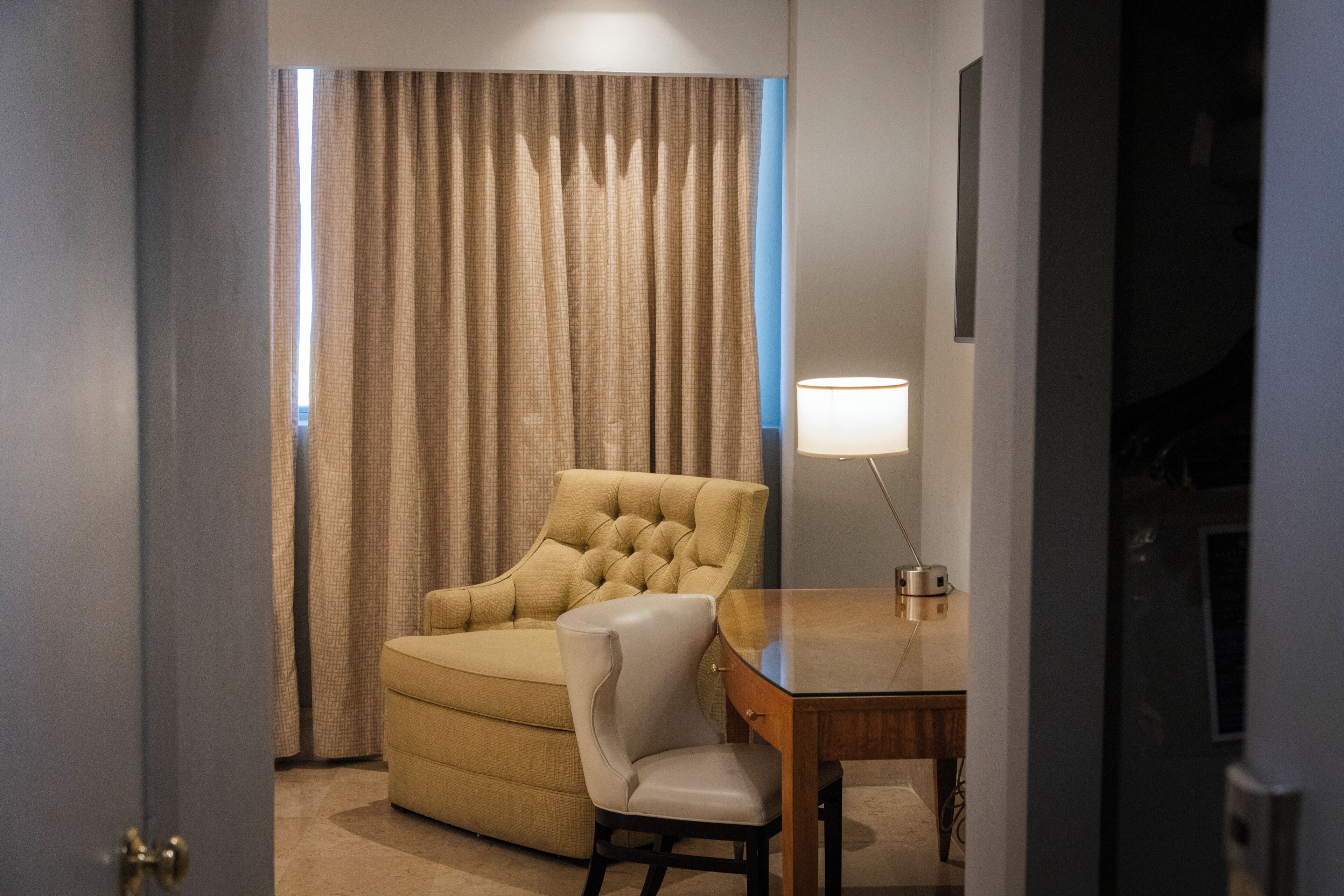 A room with a tufted armchair, a small table with a lamp, a curtain, and a door.