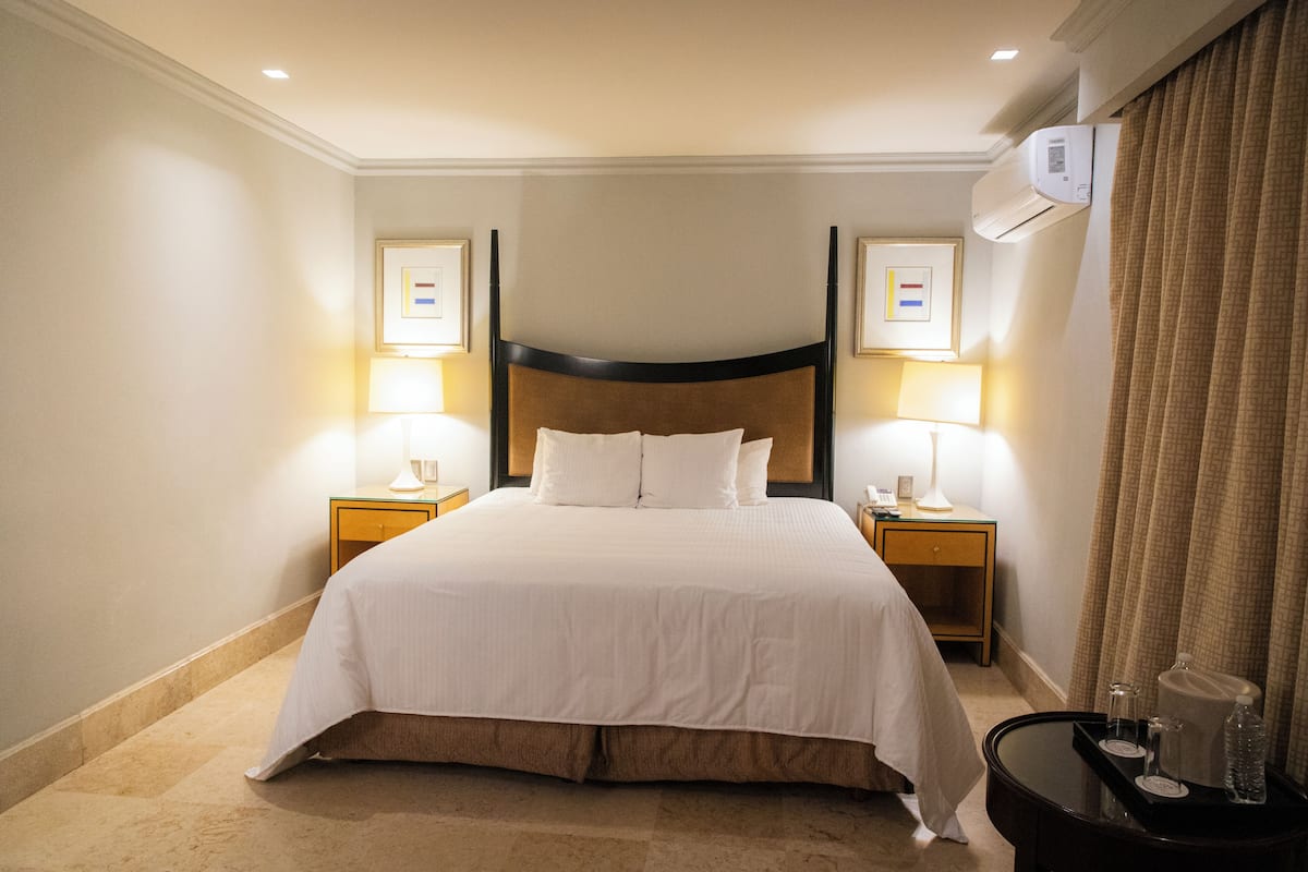 A hotel room with a large bed, bedside lamps, a nightstand, and an air conditioning unit.