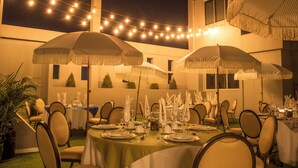 Un área de comedor al aire libre con mesas dispuestas para una comida, luces colgantes y sombrillas que brindan sombra.