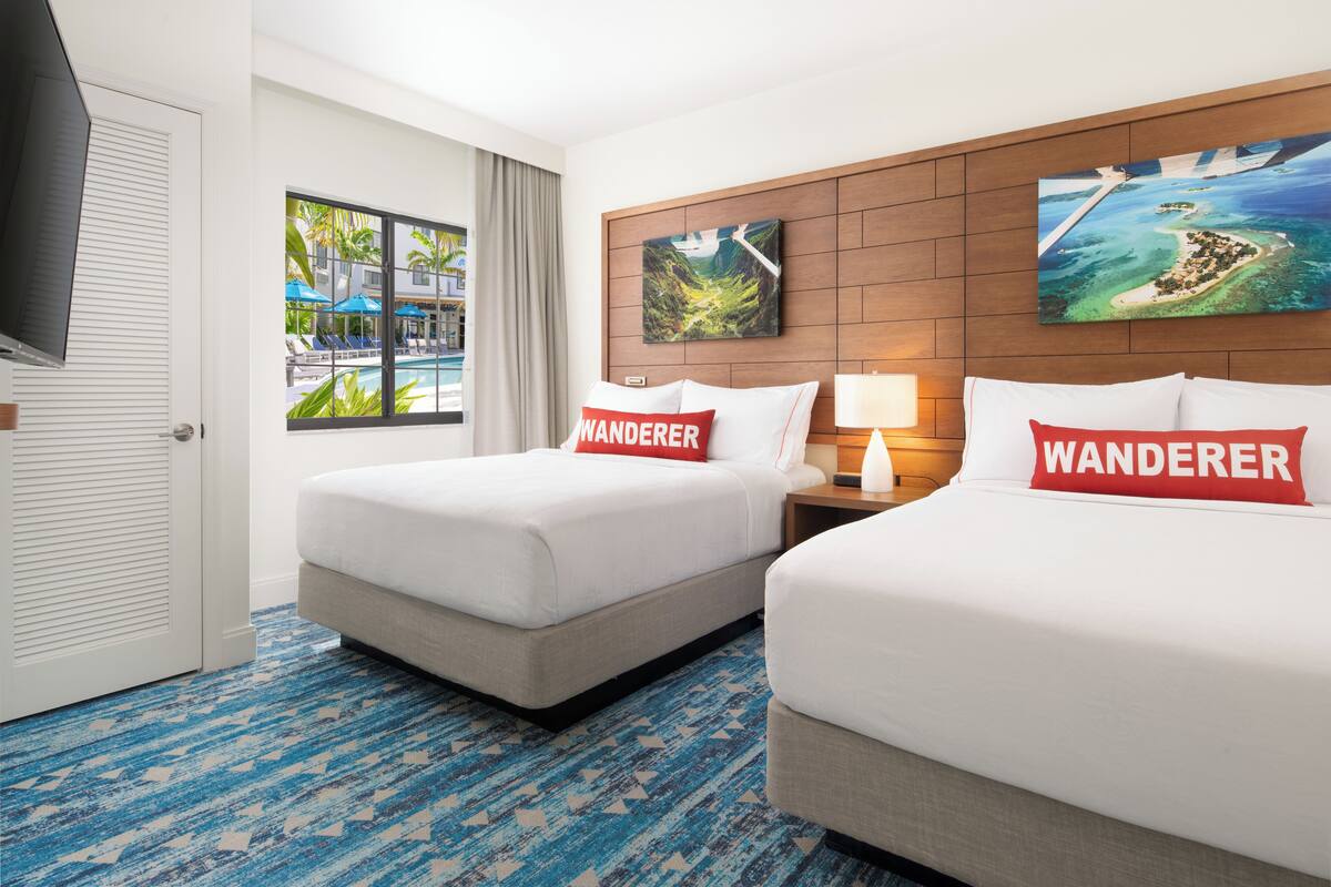A hotel room with two beds, each with a 'Wanderer' pillow, a TV, a window with a view of a pool and palm trees, and a painting of a tropical island.