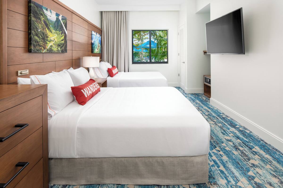 A hotel room with two beds, a wooden dresser, a TV mounted on the wall, and a window with a view of greenery.