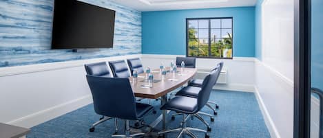 A conference room with a large table, six chairs, a flat-screen TV, and a window with a view of palm trees.