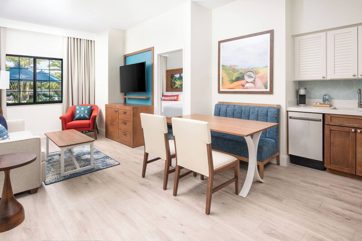 A modern living room with a wooden dining table, two chairs, a red armchair, a TV, and a kitchen area with cabinets and a countertop.