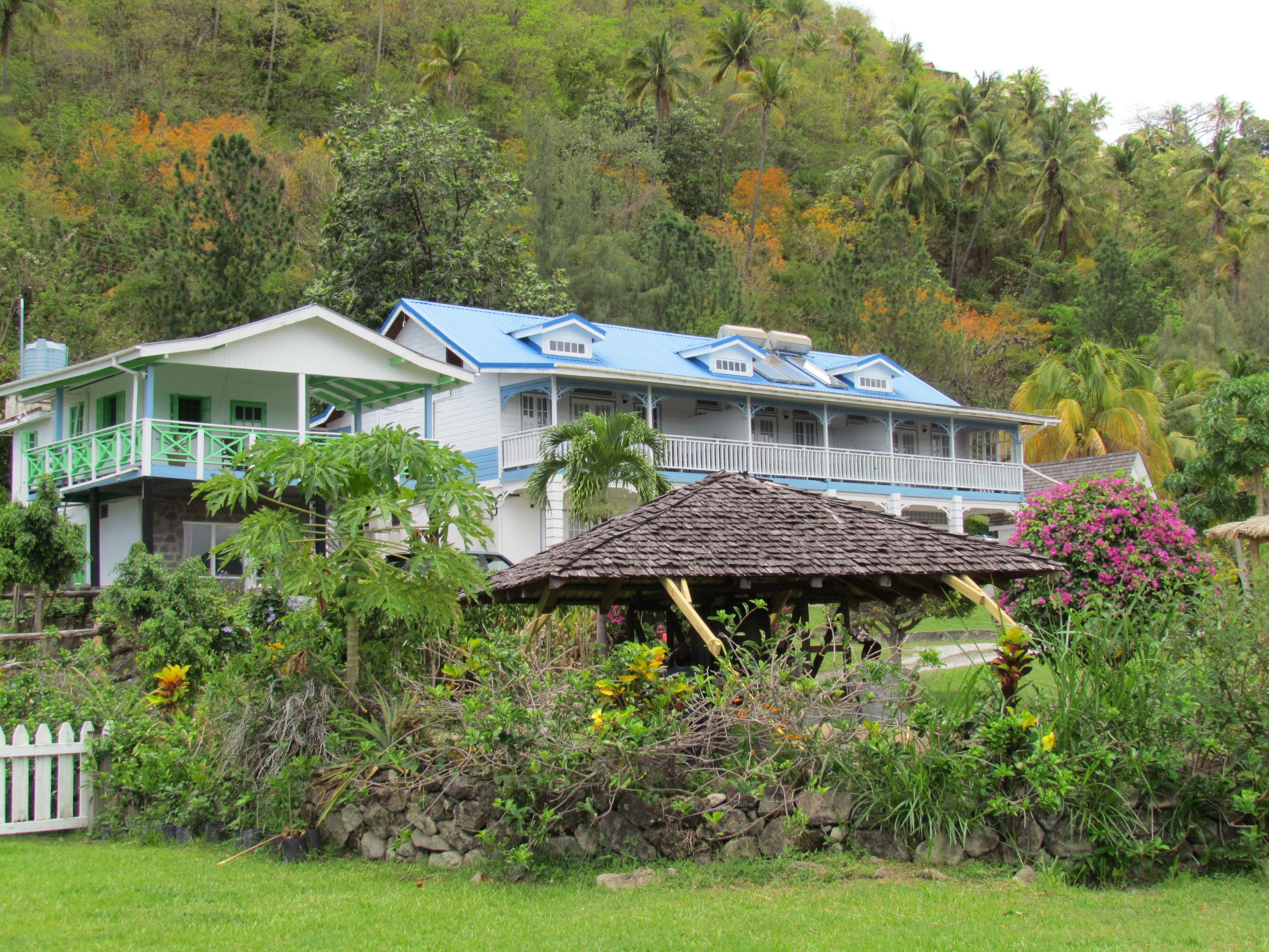 A two-story house with a blue roof and white walls, surrounded by lush greenery and a stone wall.