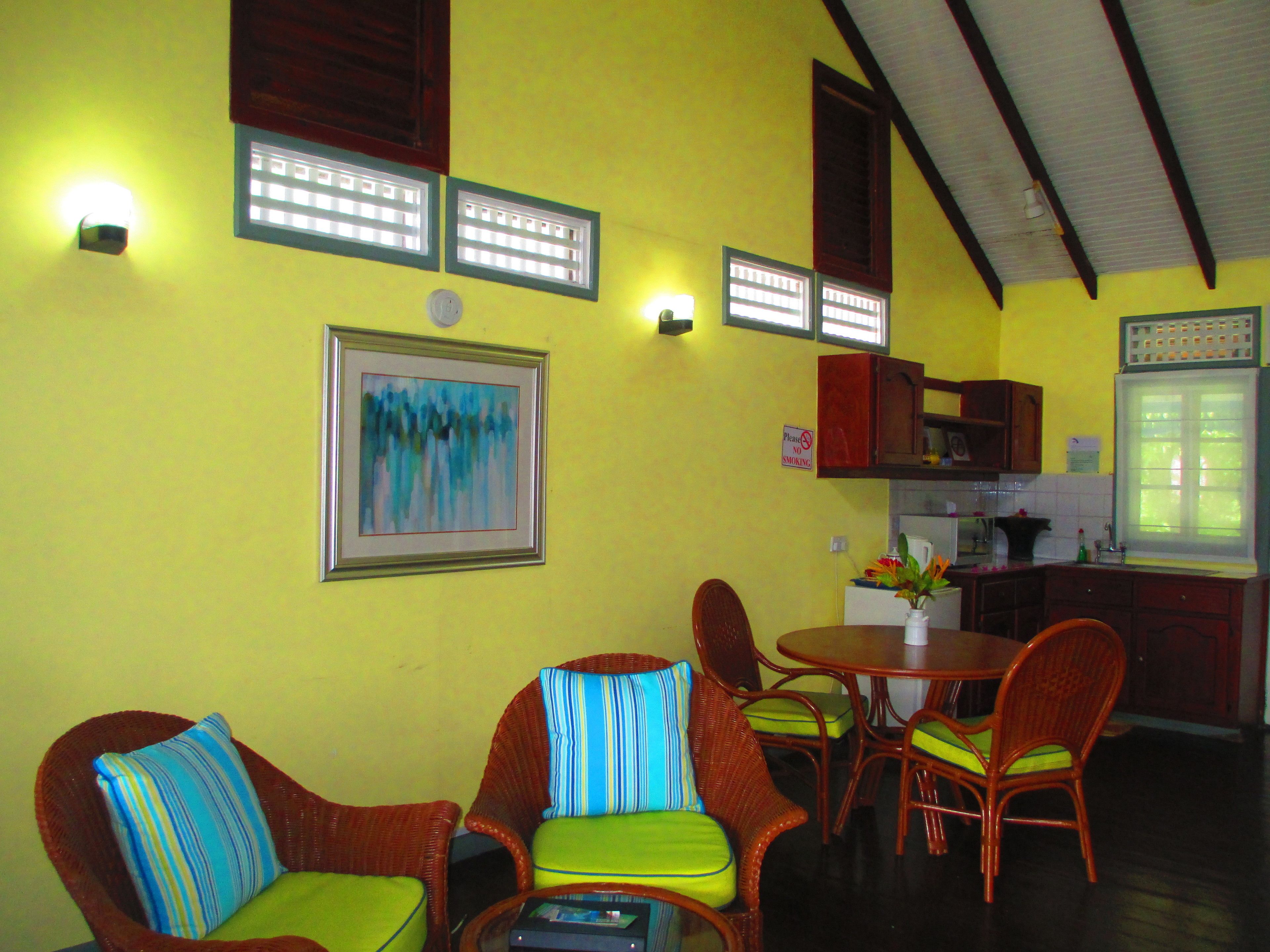 A dining area with wicker chairs and a table, a kitchen with wooden cabinets, and a yellow wall with framed artwork.