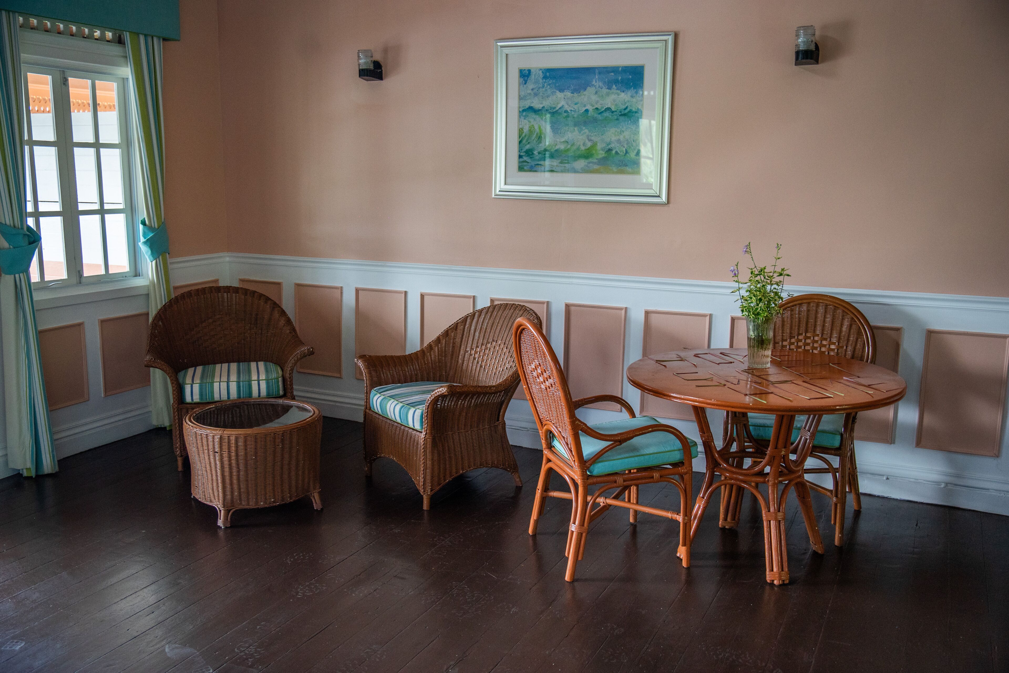 A room with a round wooden table and wicker chairs, a framed painting on the wall, and a window with curtains.
