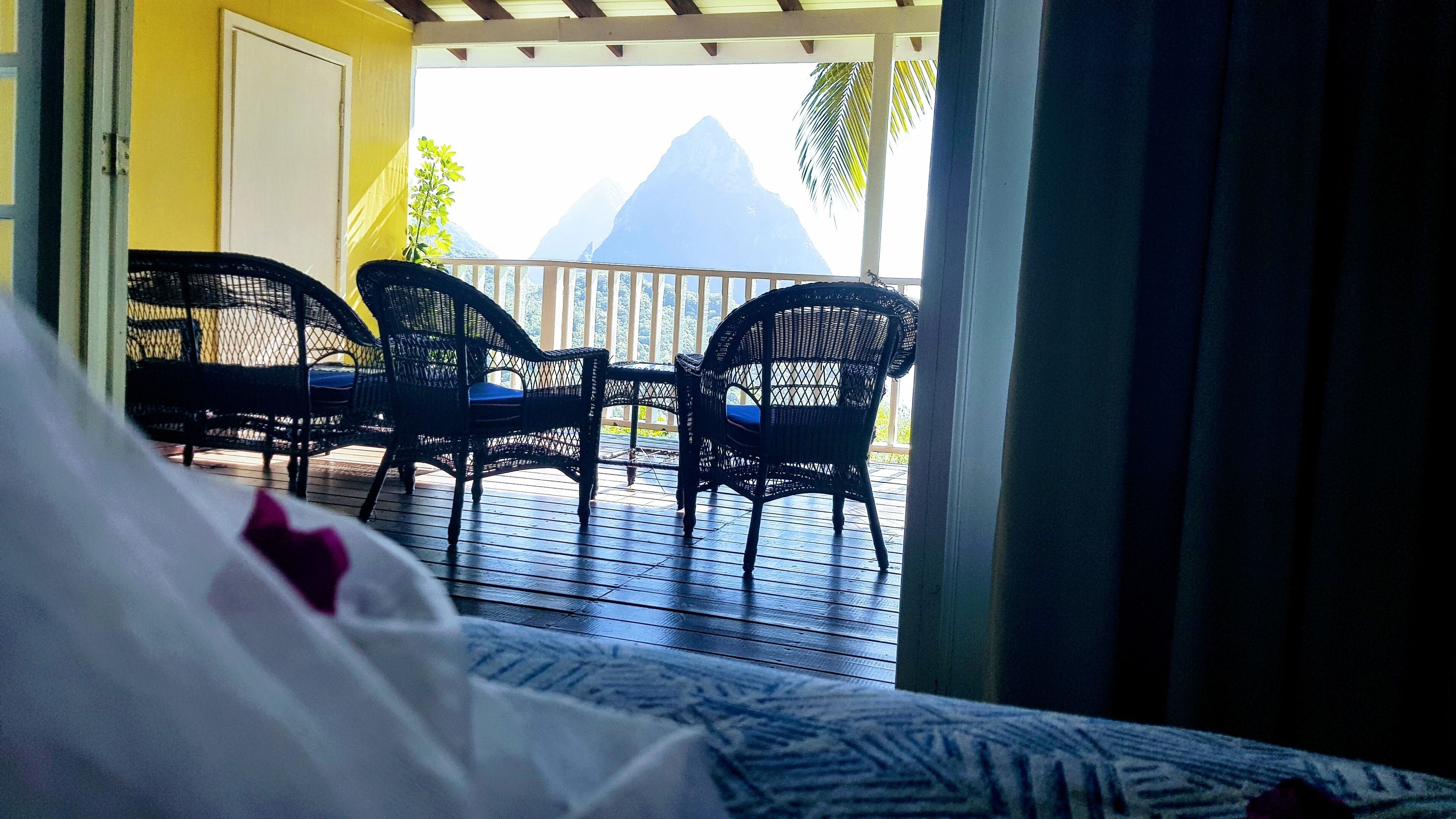 A room with a bed, two wicker chairs, and a view of mountains.
