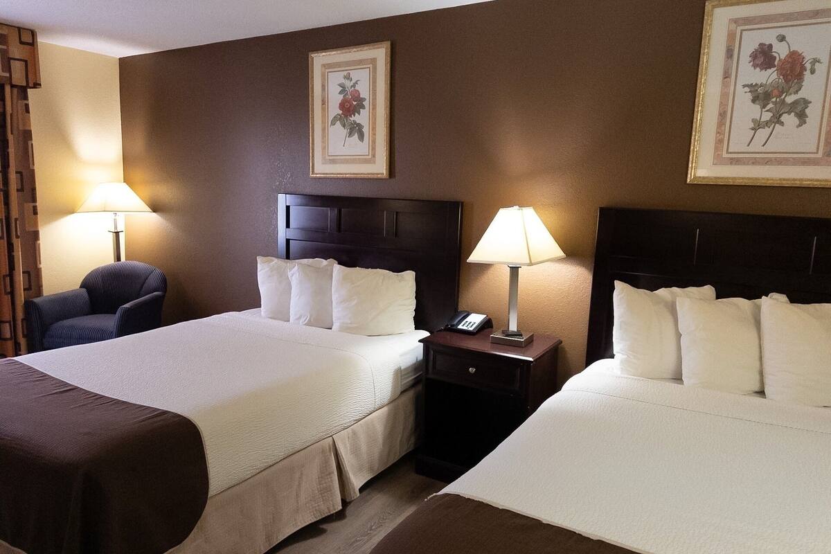 A hotel room with two beds, a chair, a nightstand with a lamp, and two framed floral pictures on the wall.