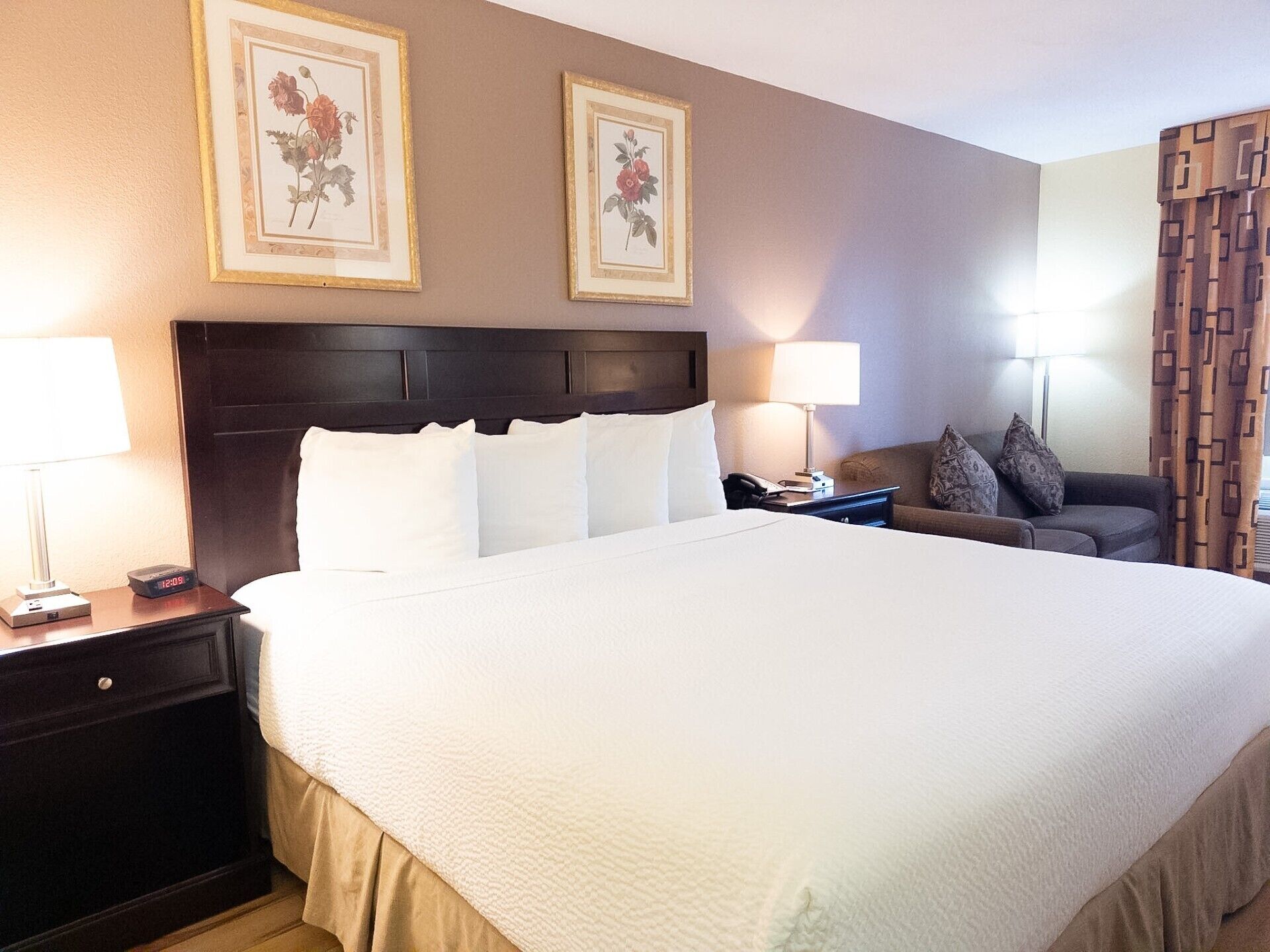 A hotel room with a large bed, two bedside tables, a headboard, a sofa, and two framed floral pictures on the wall.