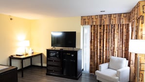 Ein Hotelzimmer mit Flachbildfernseher, Schreibtisch mit Lampe, Stuhl und gemusterten Vorhängen.