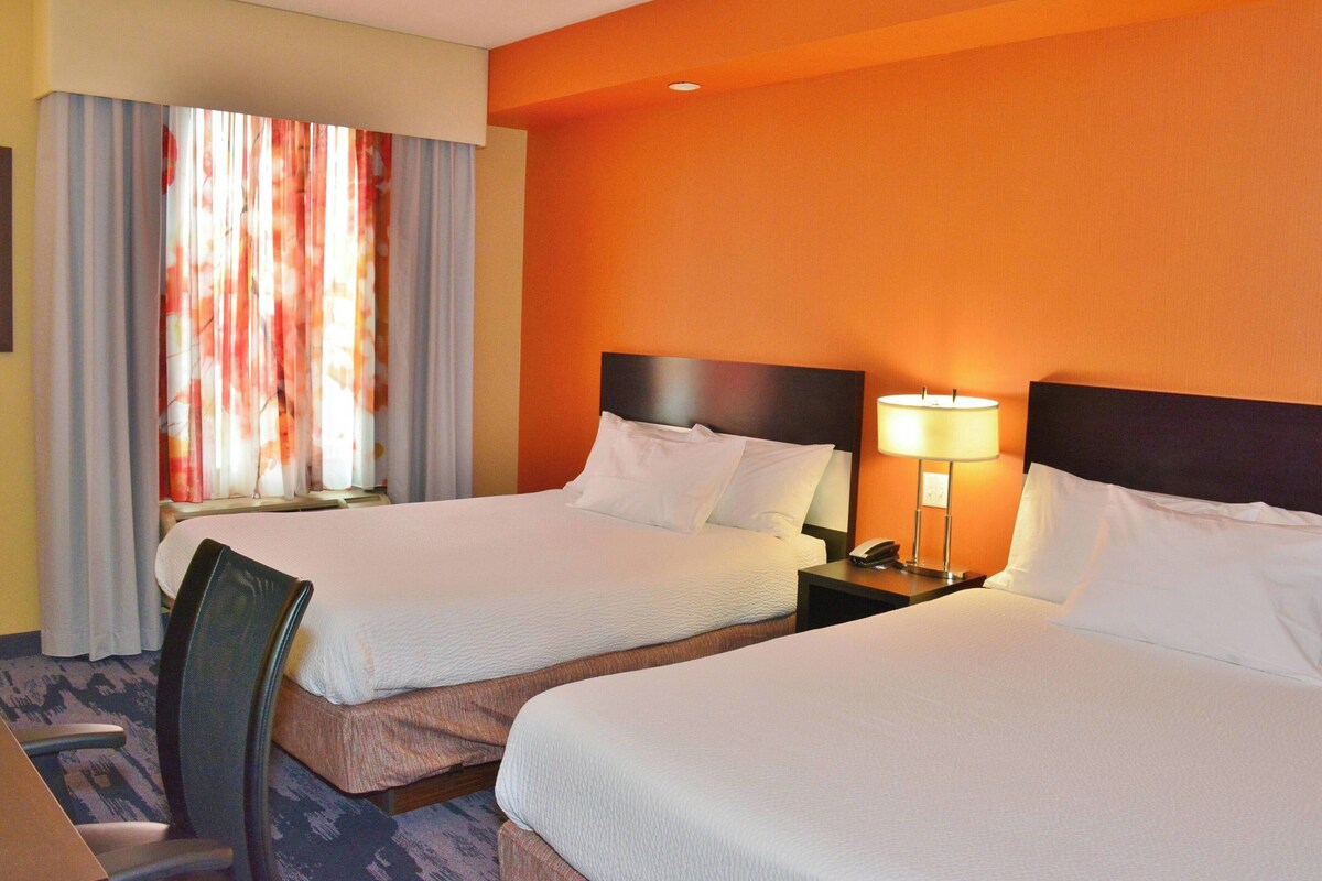 A hotel room with two beds, a chair, a nightstand with a lamp, and a window with curtains.