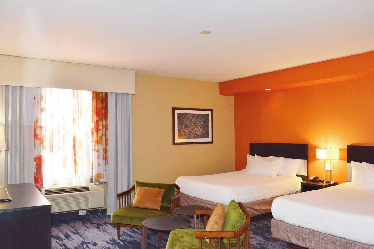 A hotel room with two beds, a desk, a chair, a small table, and a window with curtains.
