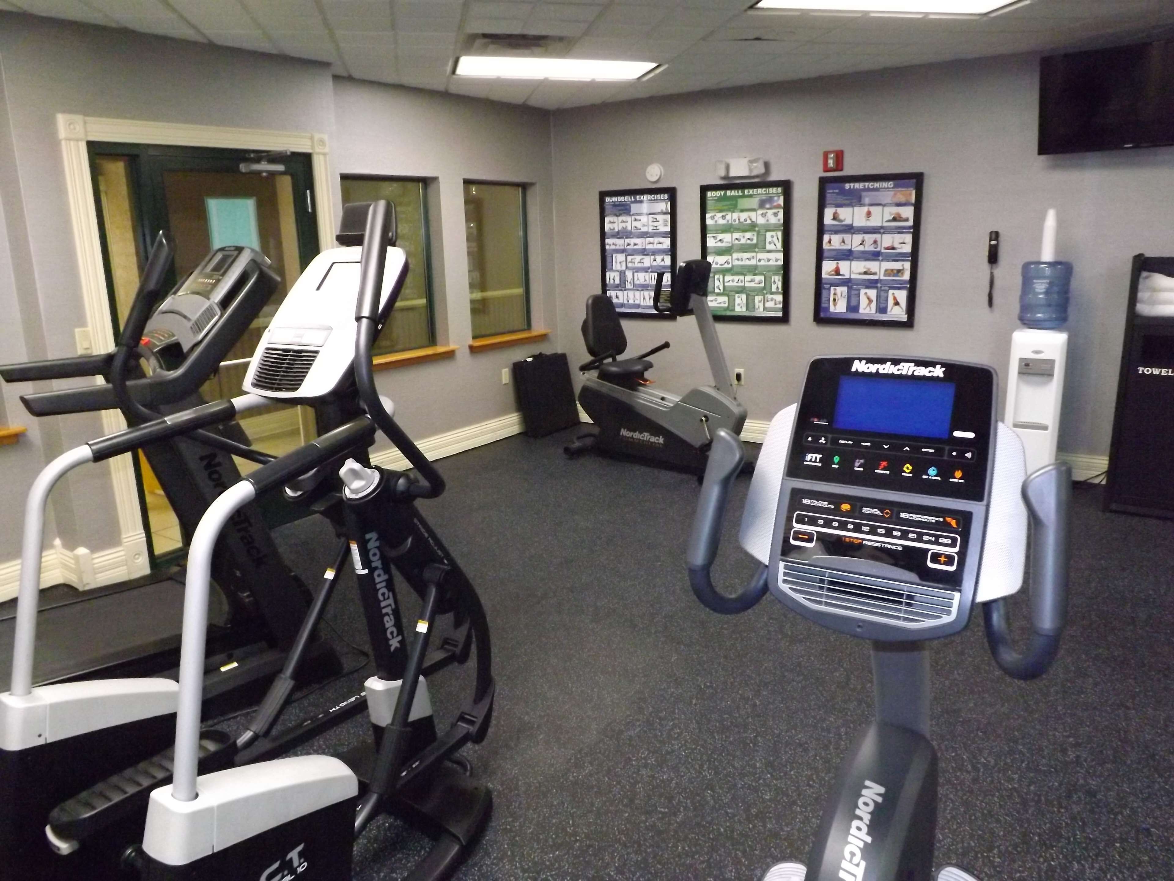 A gym with NordicTrack elliptical machines, a stationary bike, and a treadmill. There are exercise posters on the wall and a water dispenser.