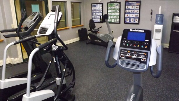 Ein Fitnessraum mit NordicTrack Crosstrainern, einem Ergometer und einem Laufband. An den Wänden befinden sich Trainingsposter und ein Wasserspender.