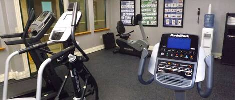A gym with NordicTrack elliptical machines, a stationary bike, and a treadmill. There are exercise posters on the wall and a water dispenser.