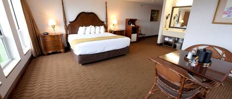 A hotel room with a large bed, a desk with a chair, a dresser, a vanity with a mirror, and a window with curtains.