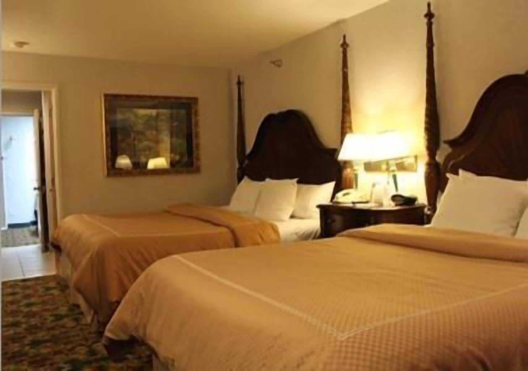 Quarto de hotel com duas camas, uma mesa de cabeceira com abajur, um quadro na parede e uma porta que dá para outro cômodo.