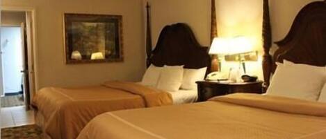 Quarto de hotel com duas camas, uma mesa de cabeceira com abajur, um quadro na parede e uma porta que dá para outro cômodo.