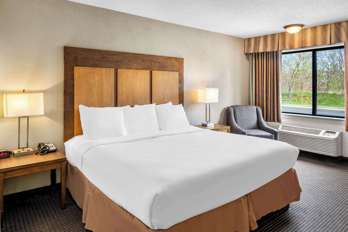 A hotel room with a large bed, two bedside lamps, a chair, a desk with a phone, and a window with curtains.