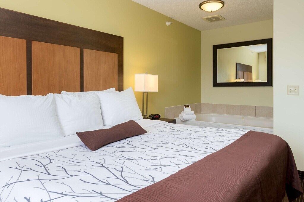 Una habitación de hotel con una cama grande, cabecera de madera, mesita de noche con lámpara, baño con espejo y perchero para toallas.