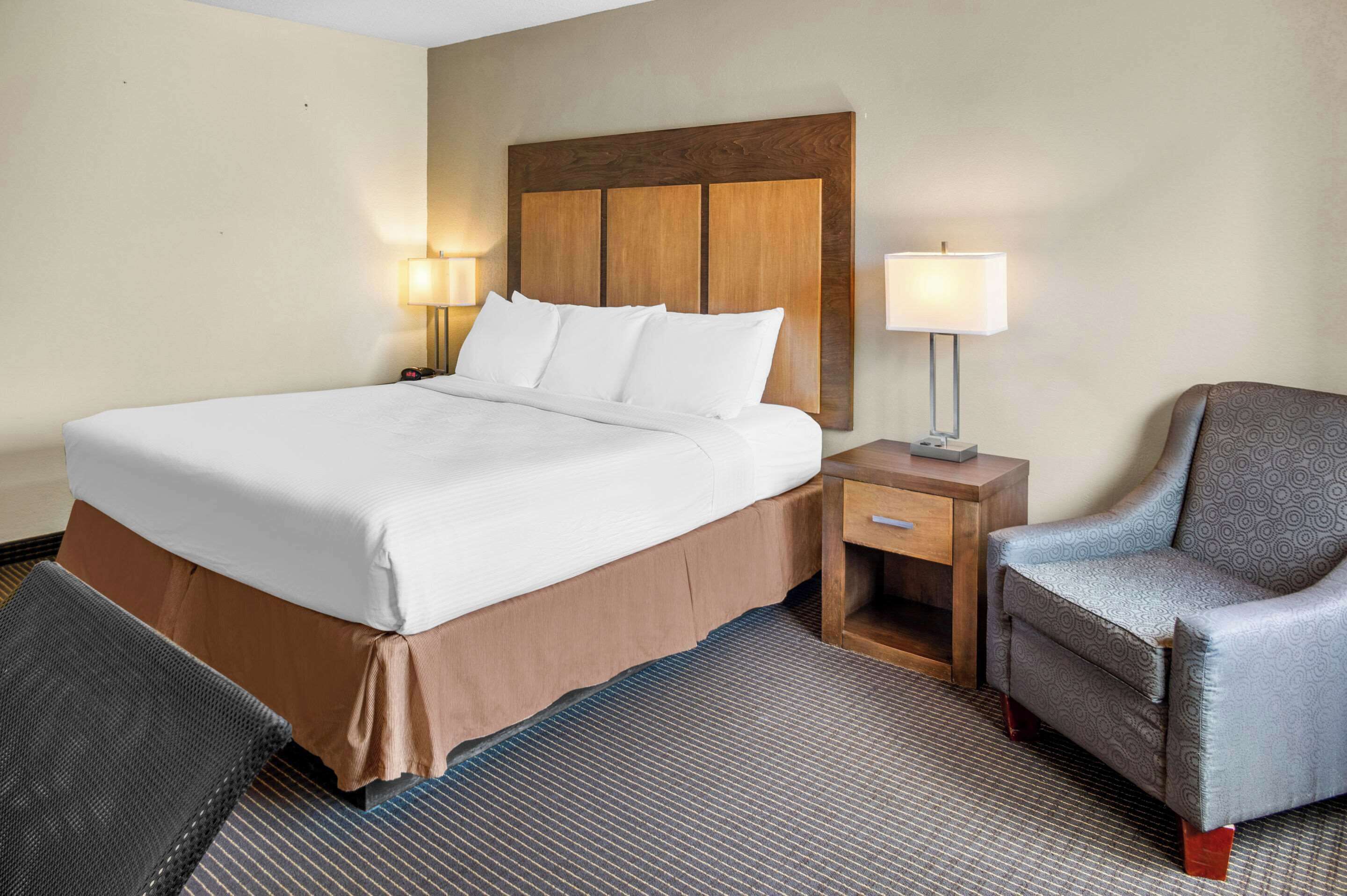 Una habitación de hotel con una cama grande, dos mesitas de noche con lámparas, una silla y piso alfombrado.