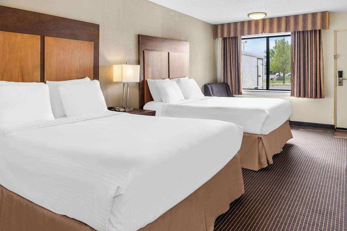 A hotel room with two beds, a nightstand, a lamp, a chair, and a window with curtains.