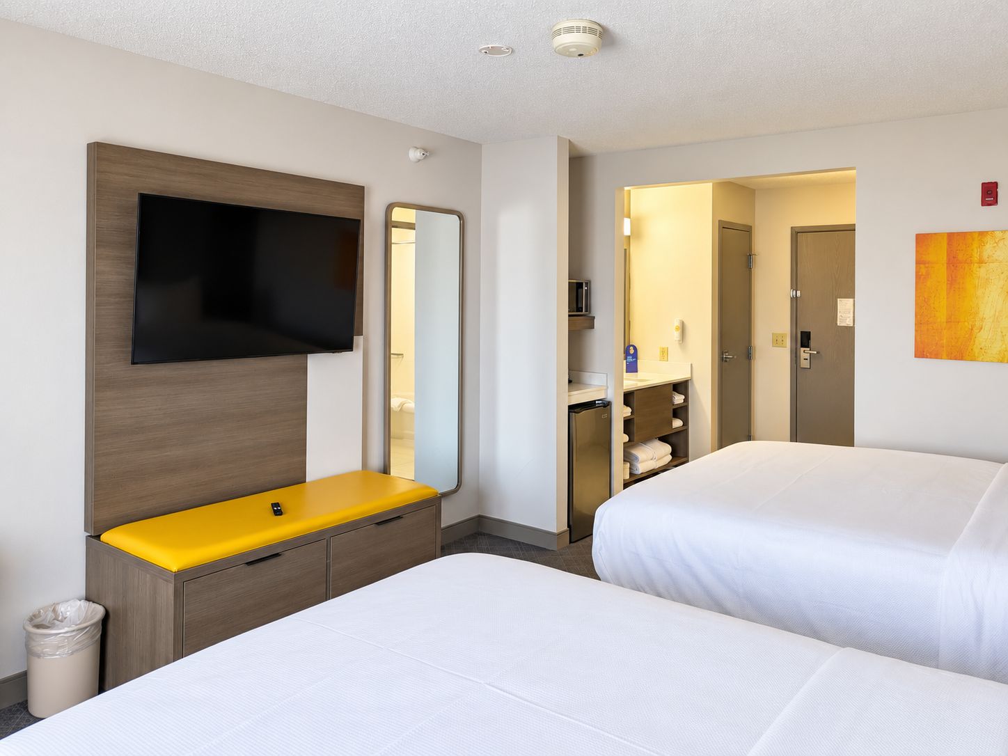 A hotel room with a bed, a TV mounted on the wall, a mirror, a small desk with a yellow surface, and a door leading to a bathroom.