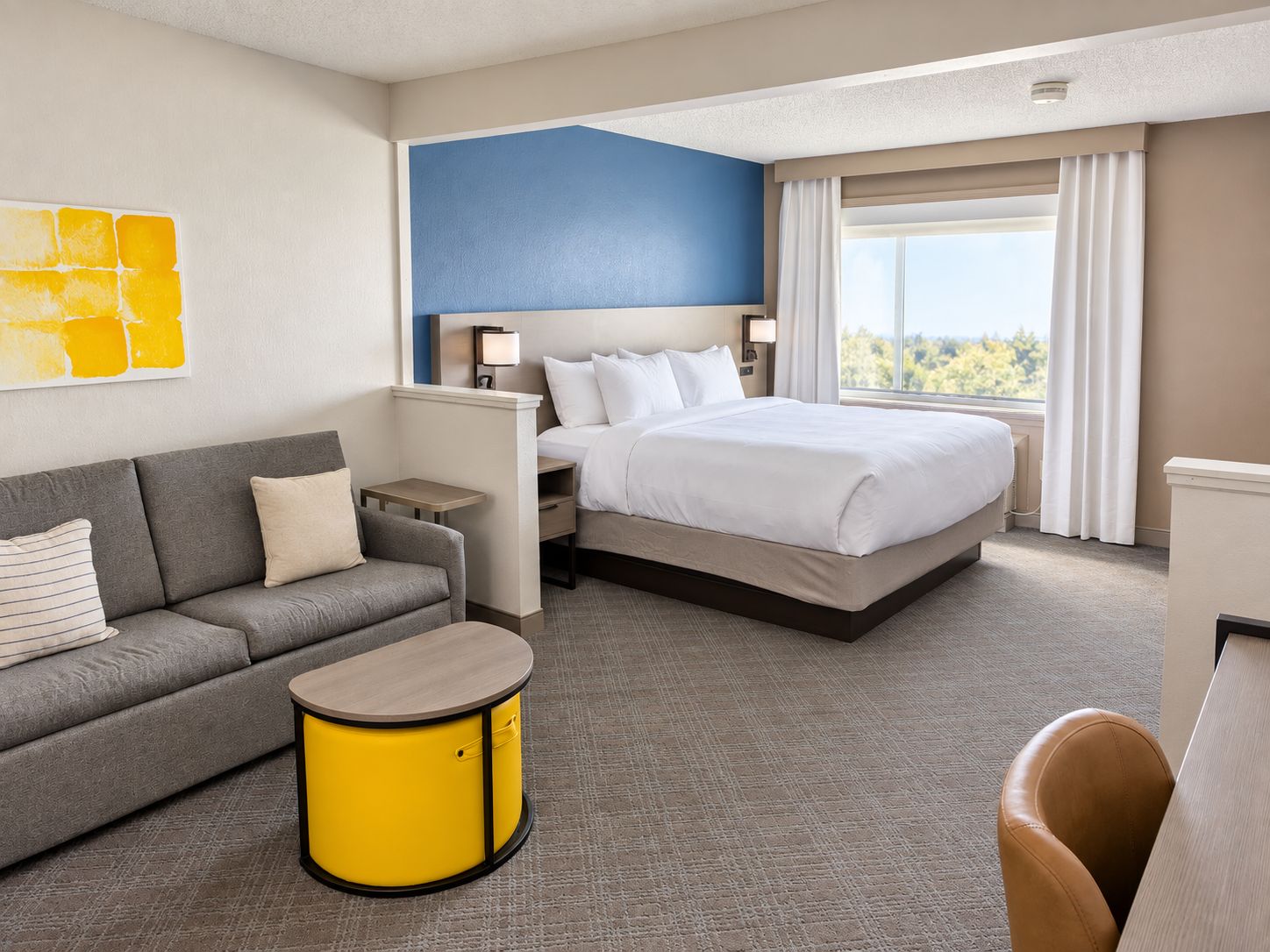 Quarto de hotel com cama, sofá, mesa pequena e mesa lateral amarela.