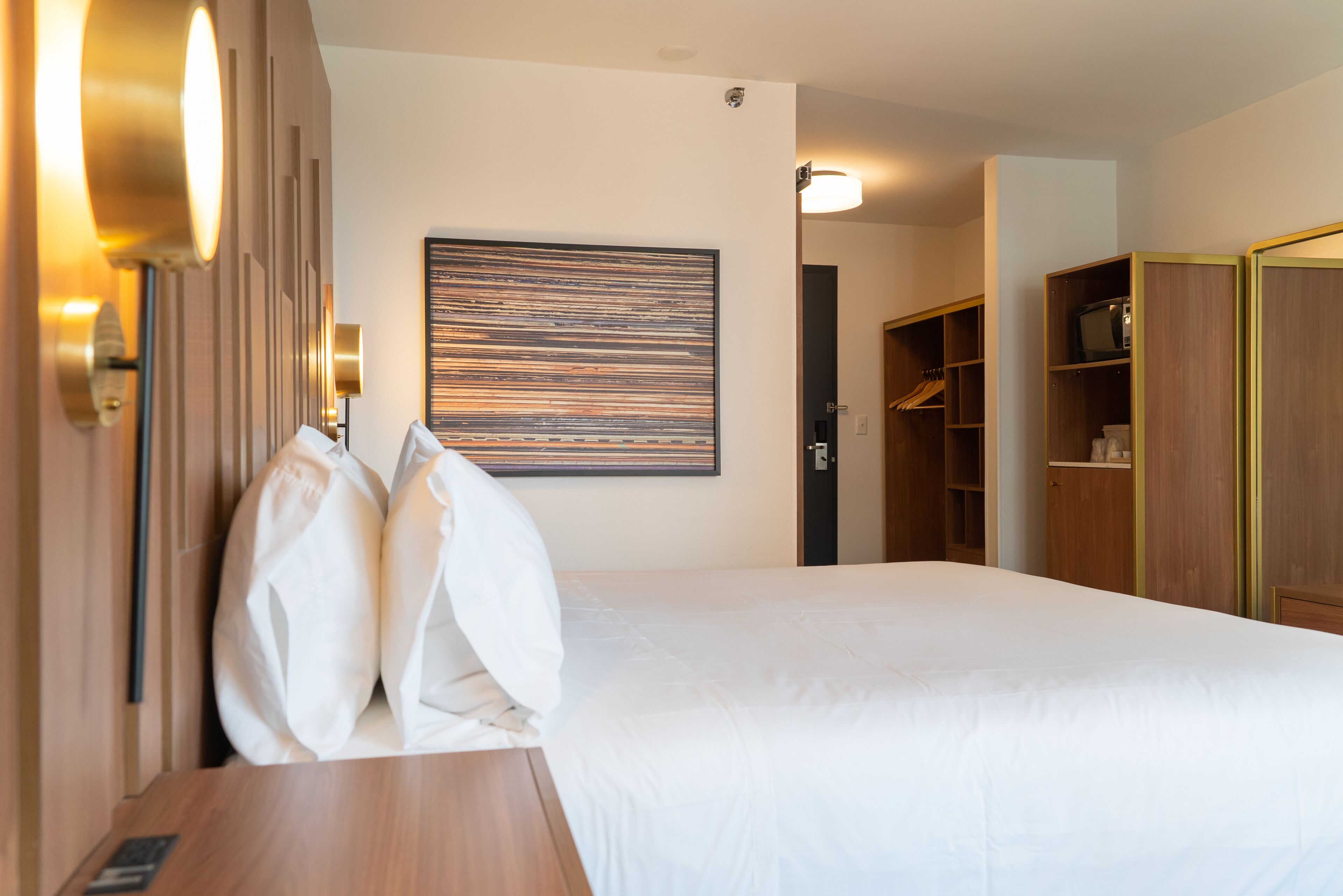 A hotel room with a bed, a bedside table, a wall-mounted artwork, and built-in wooden shelving.